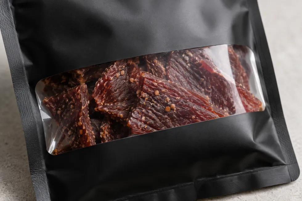 Close-up of a matte black pouch with a clear window showing dark reddish-brown beef jerky strips with visible seasoning and a textured surface.